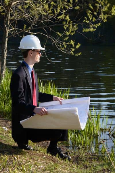 Engineer reviewing detention pond design plans - ALTA SURVEY Texas Civil engineer wearing a hard hat reviewing detention pond design plans beside a stormwater pond