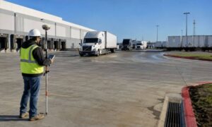 Commercial property surveyor using GPS equipment to assess a truck terminal site with semi-trucks, loading docks, and trailer circulation areas